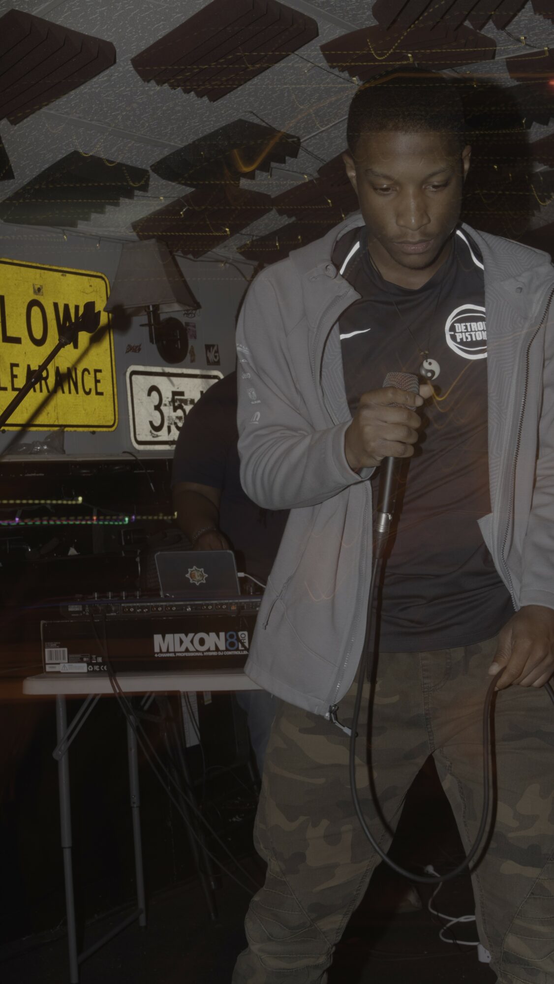 Young person holding a microphone, standing near a keyboard, with a yellow 'Slow' sign and scattered objects in background.