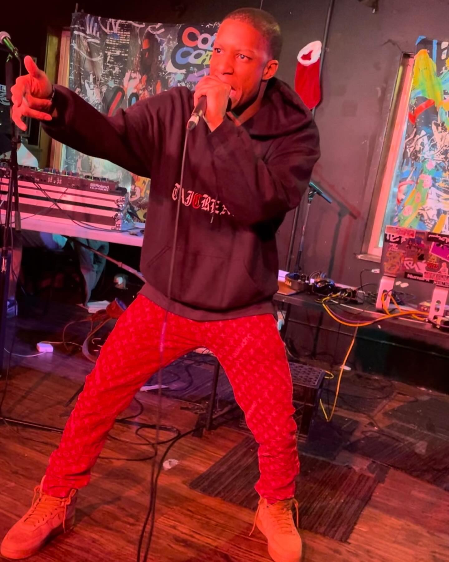 Person performing on stage with microphone, wearing black hoodie and red pants, in a colorful setting.