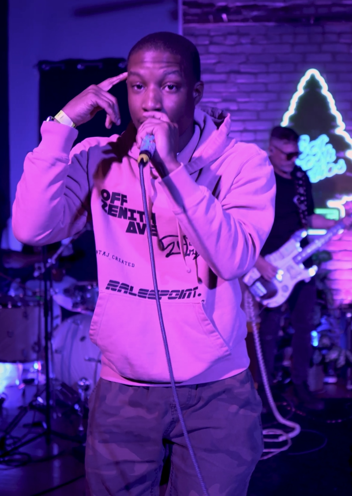 Young man singing into a microphone on stage with band, purple lighting, brick wall background, neon sign, casual clothing.