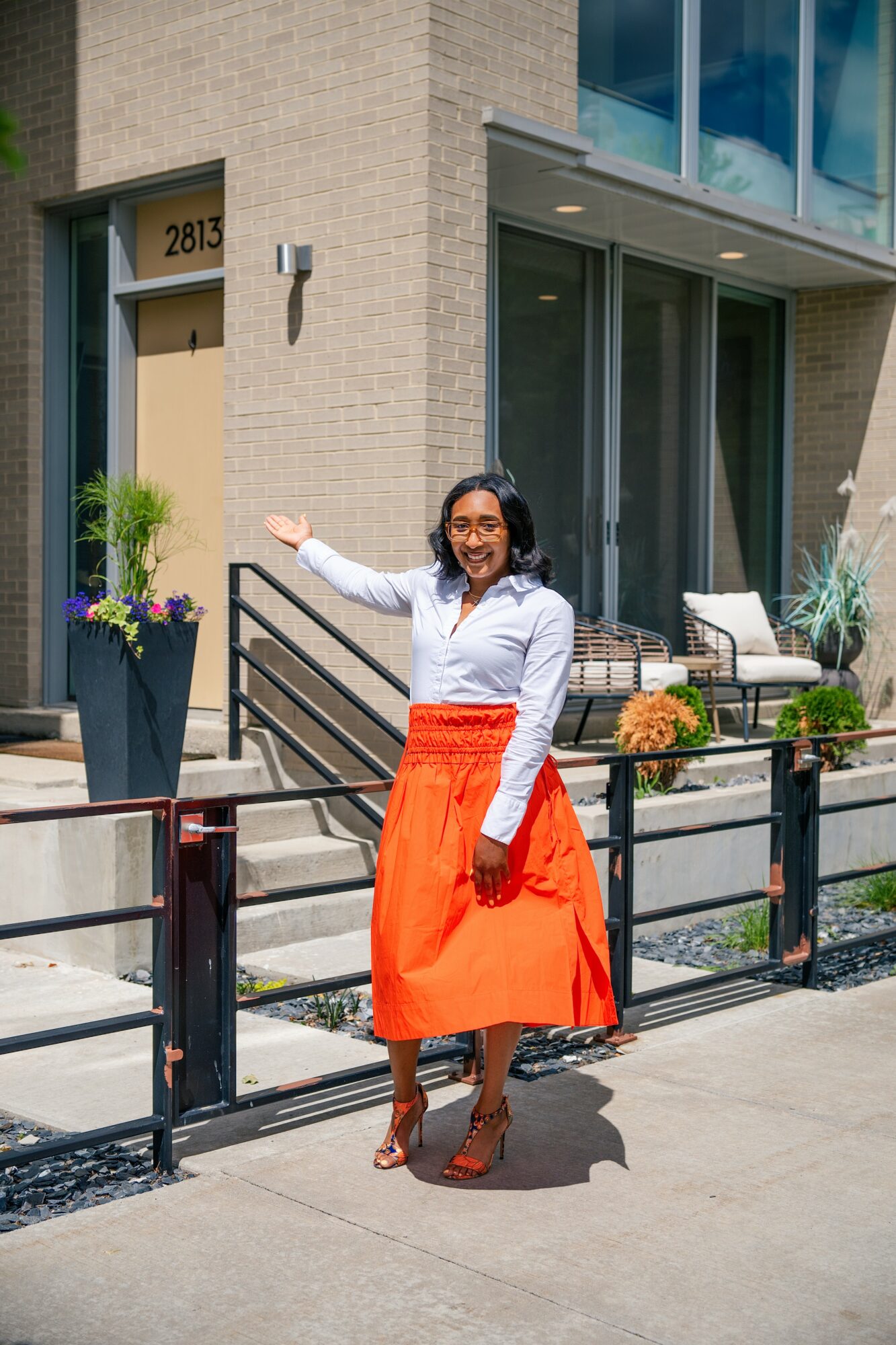 Woman in white top and orange skirt standing outside a building, smiling, with plants and seating nearby.