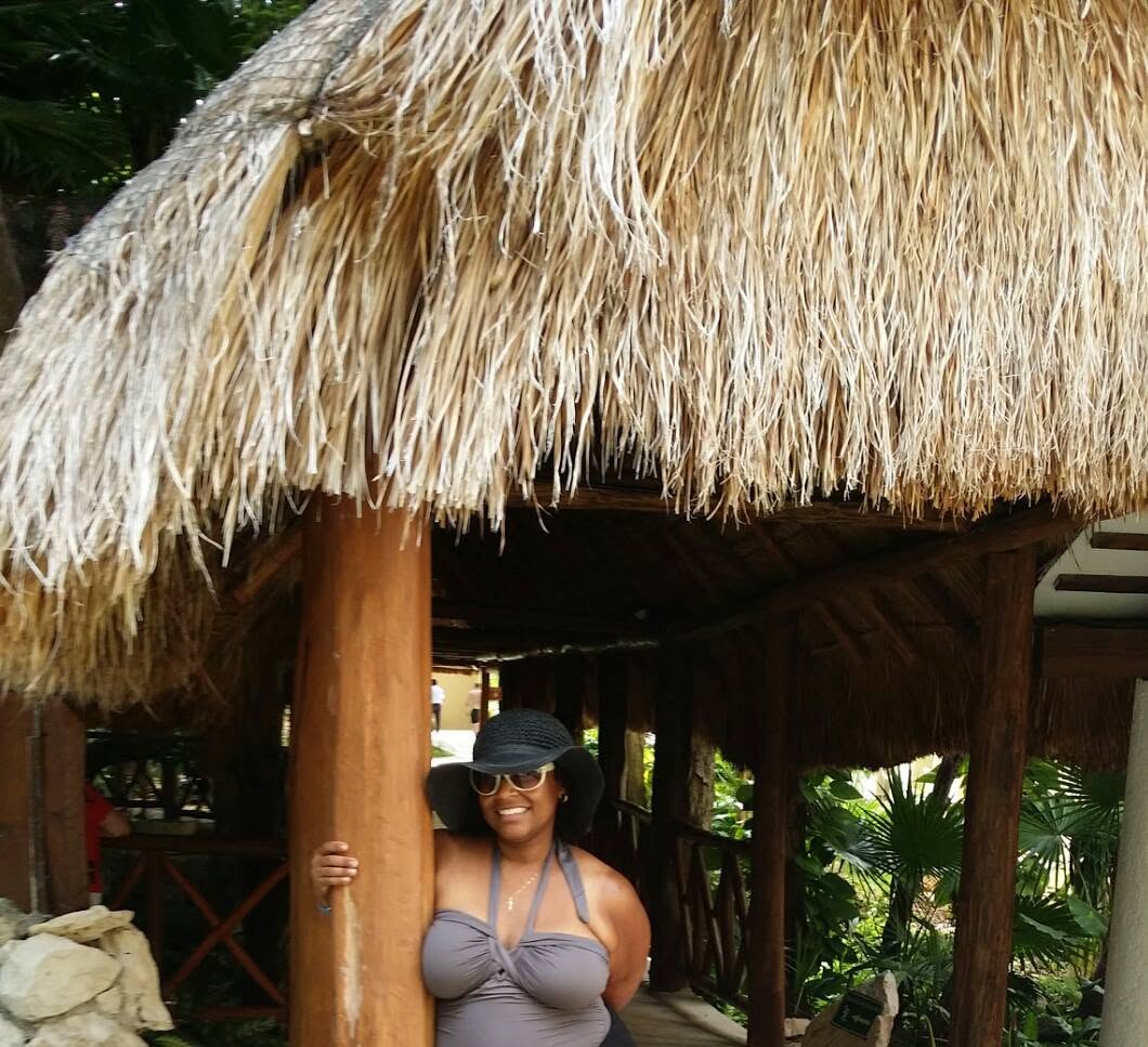 Woman in sunglasses and hat standing under a thatched roof structure outdoors, smiling, with greenery in background.