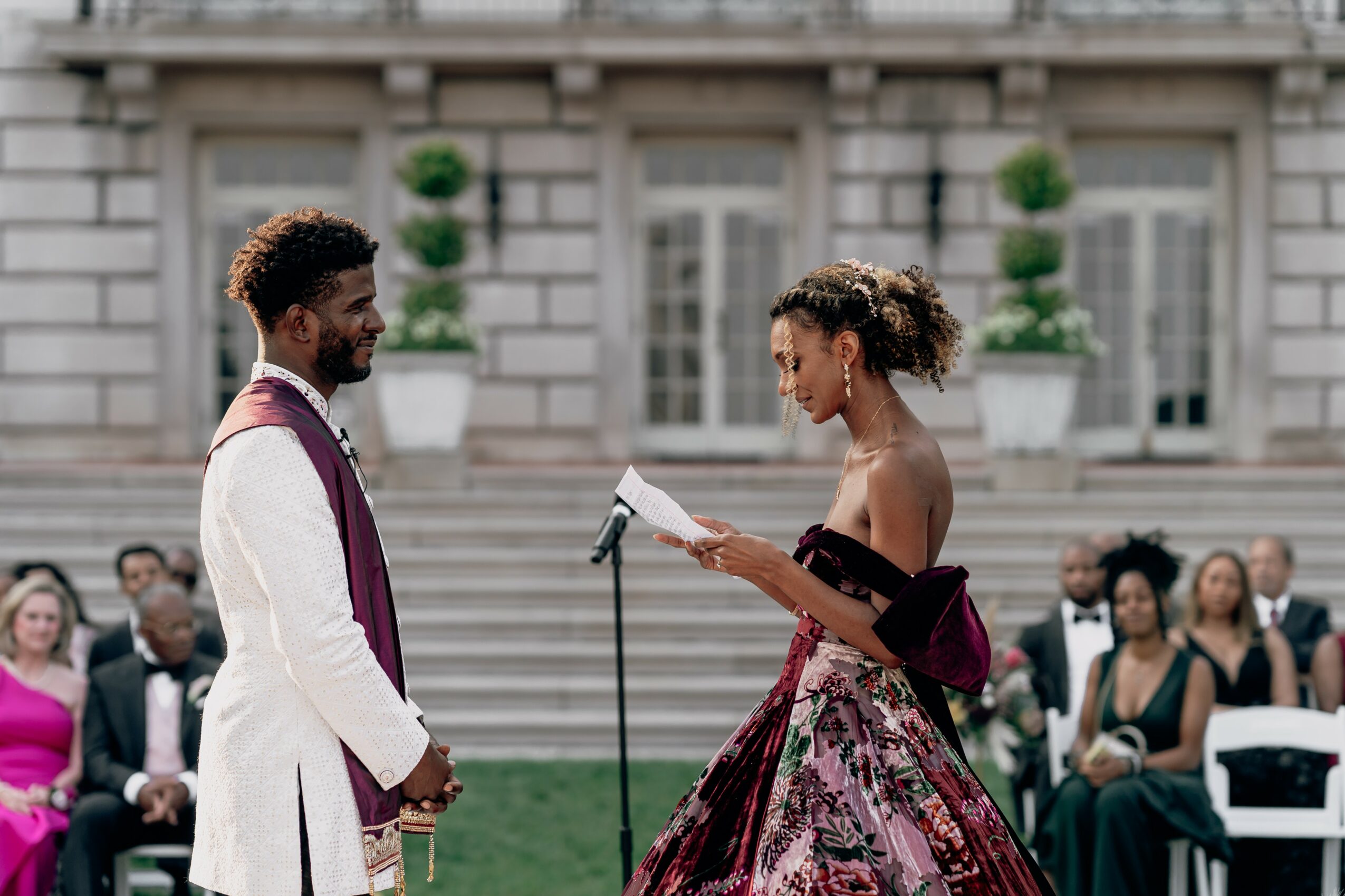 A couple stands outdoors in front of a building, with an audience seated behind them, during a formal event.