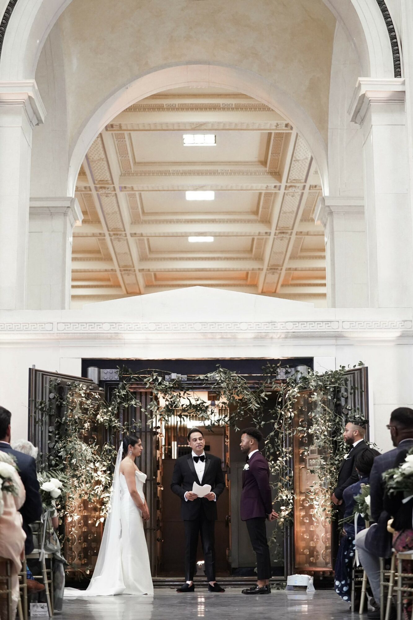 Wedding ceremony with a bride, groom, and officiant under an arch decorated with flowers, inside a grand hall.