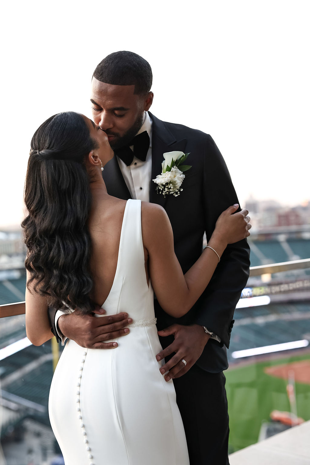 A couple in wedding attire embracing outdoors with a stadium in the background.