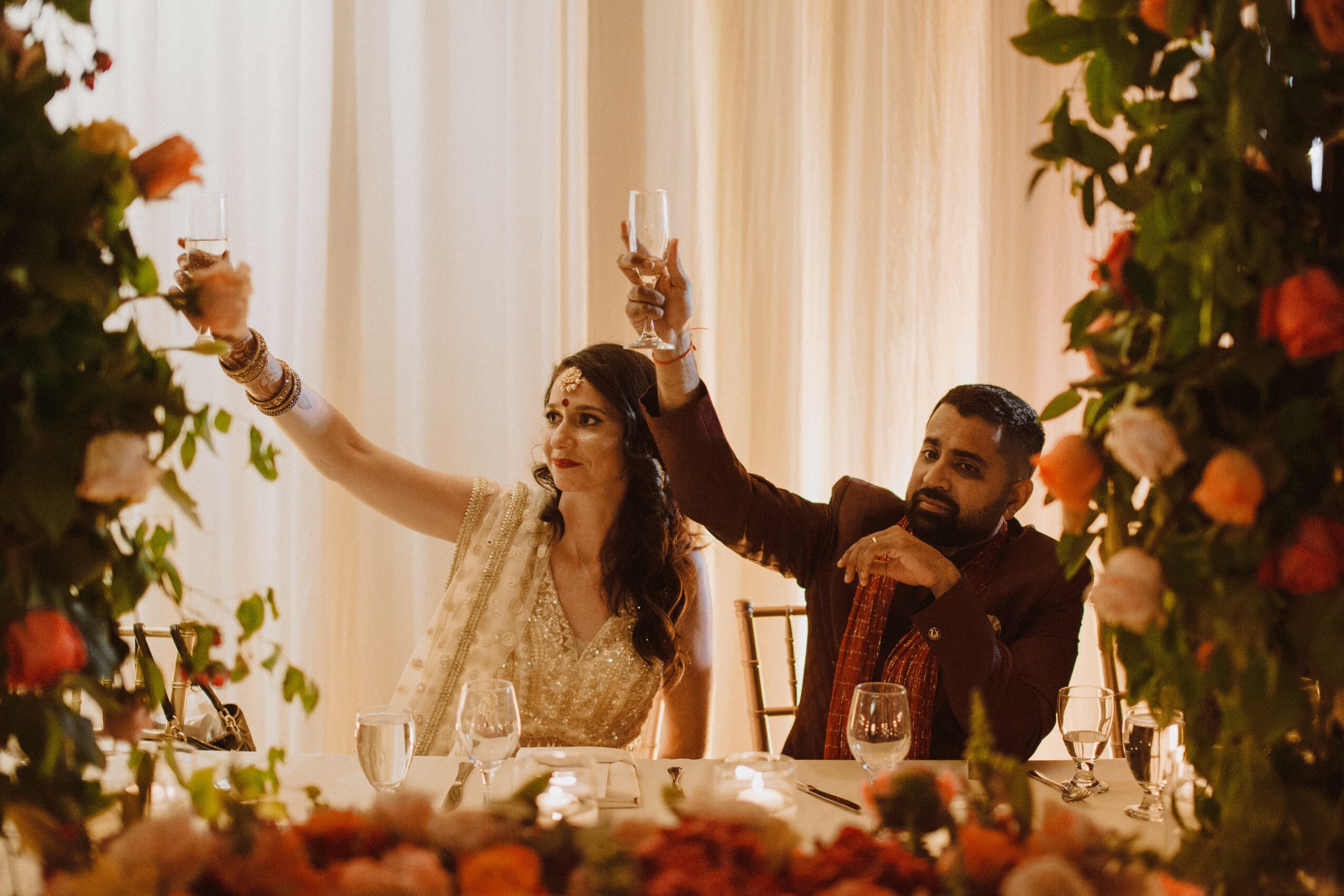 A couple at a celebration raising glasses, surrounded by floral decorations and warm lighting, smiling and enjoying the moment.