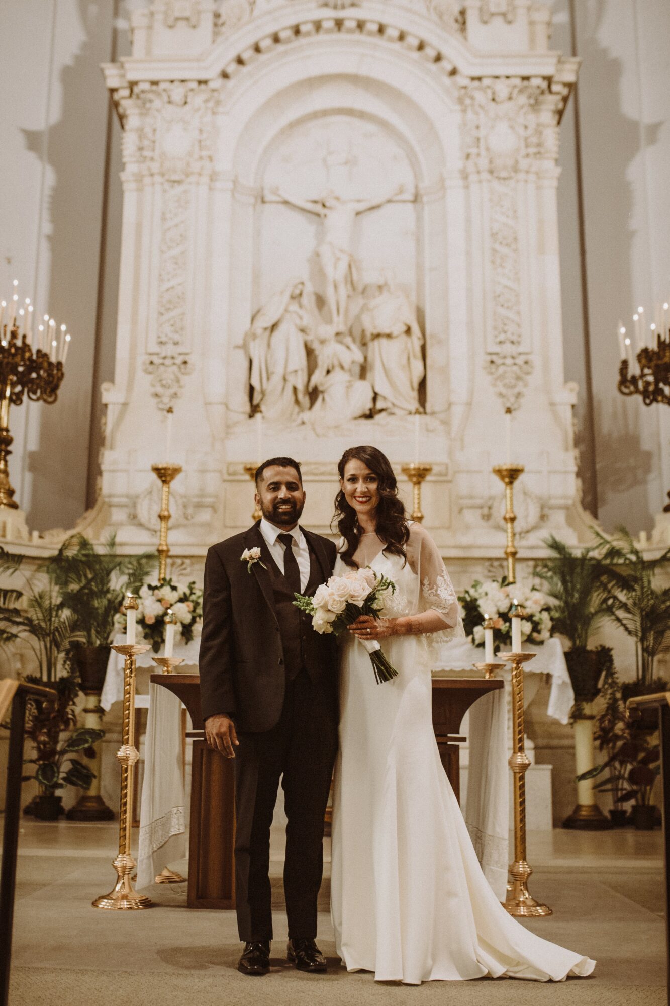 A bride and groom stand together inside a church with ornate architecture and a large religious sculpture behind them.