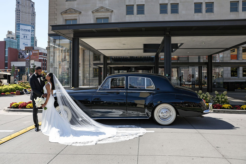 Bride and groom stand next to a vintage black car on city sidewalk, with modern buildings in background.