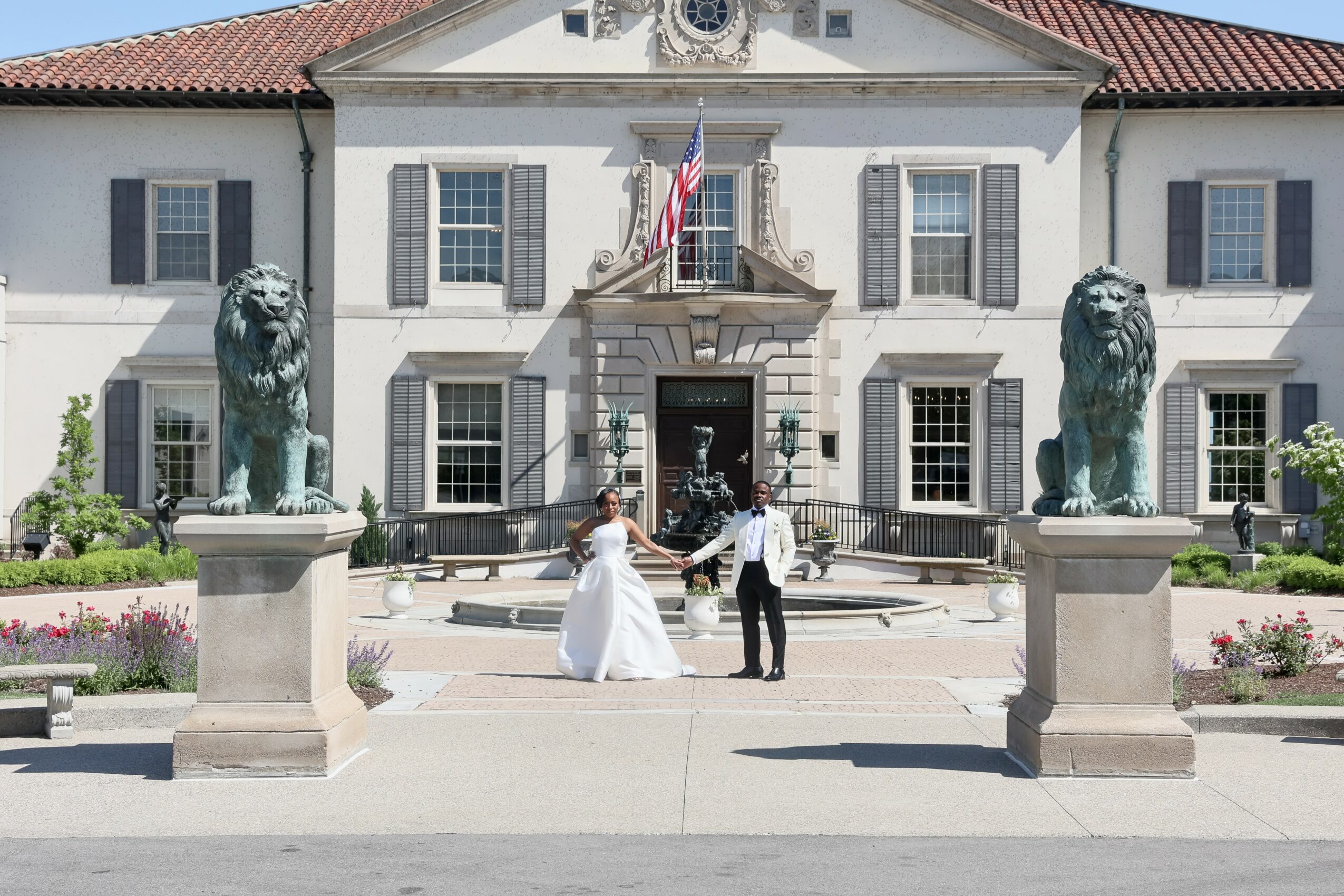 A bride and groom holding hands in front of a large white building with two lion statues on pedestals, flowers, and an American flag.