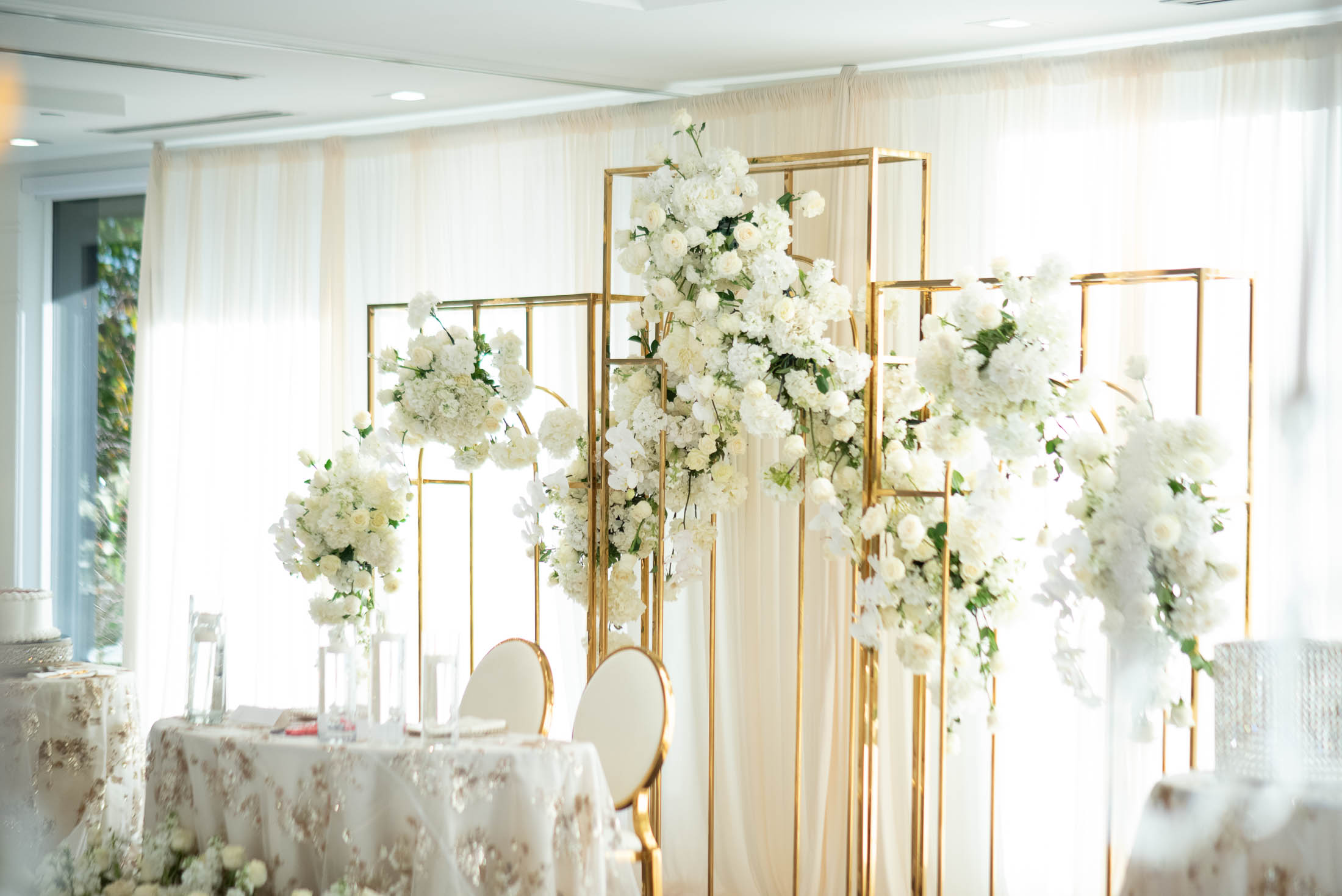 Elegant event setup with floral arrangements and a table with chairs, in a bright room with white curtains.