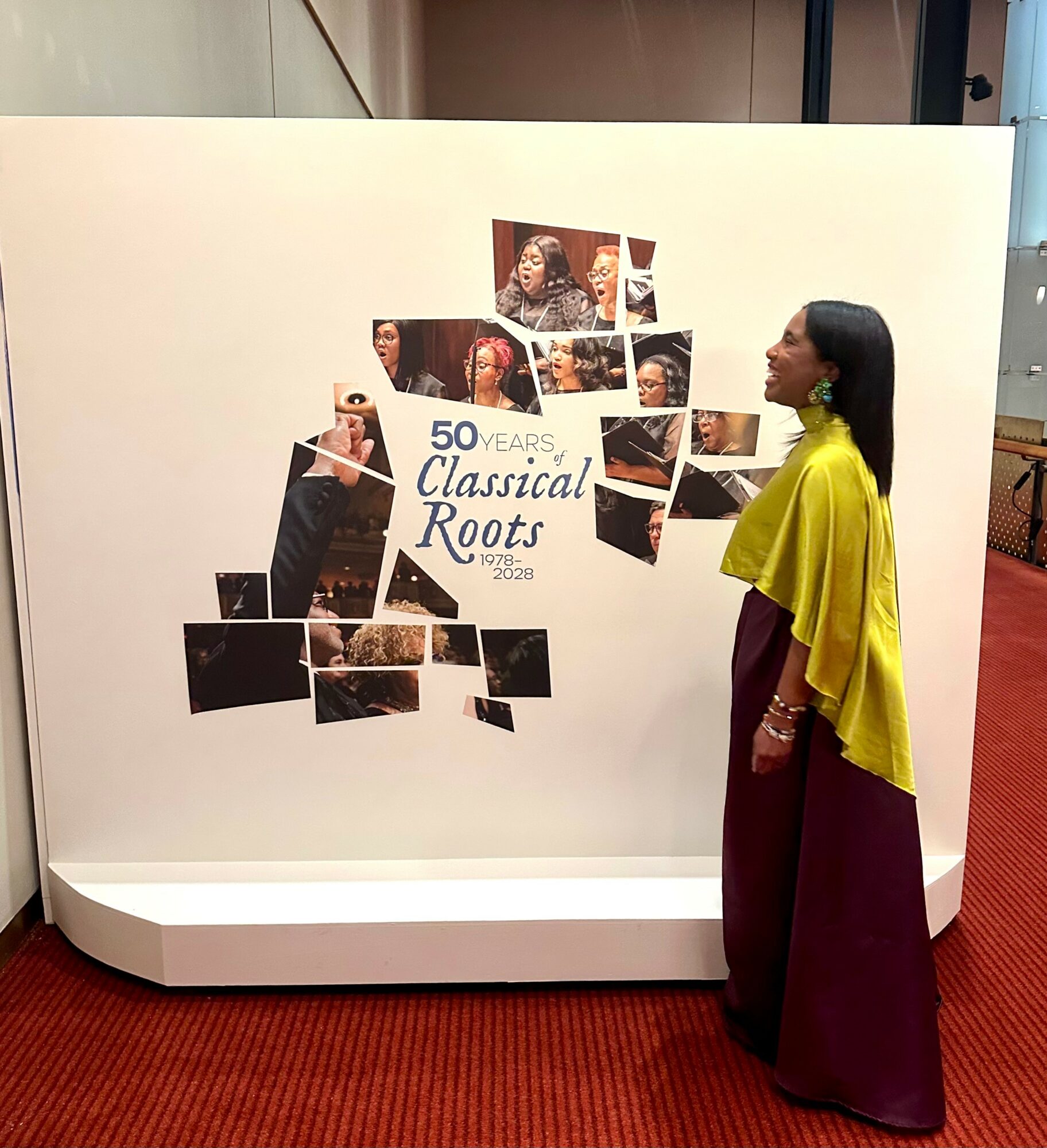 Woman in yellow and maroon dress looking at a display with photos and text about 50 years of classical roots.