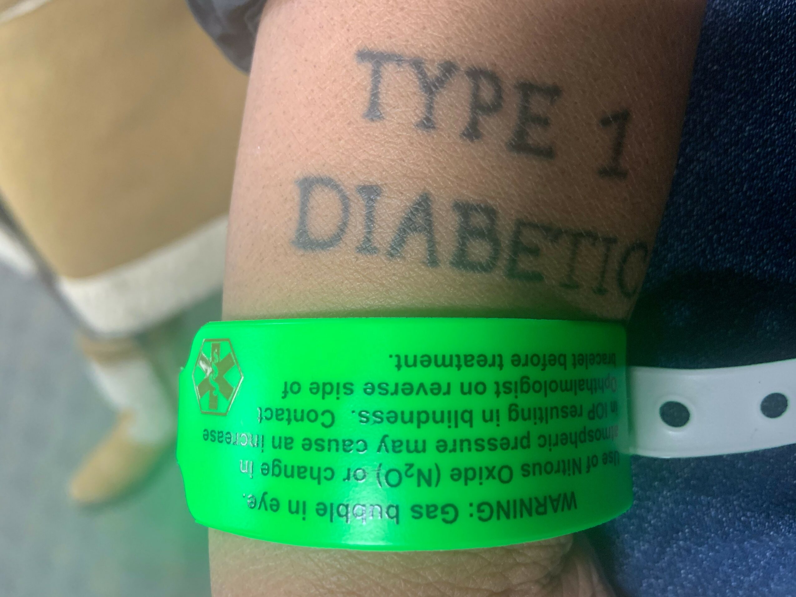 Person's arm with tattoo reading 'TYPE 1 DIABETIC' and a green allergy bracelet, close-up.