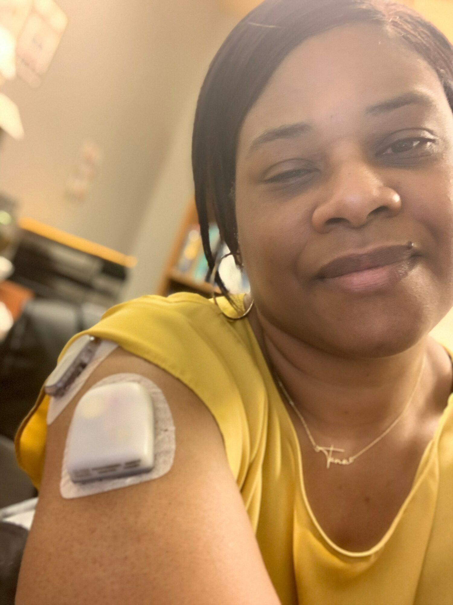 Woman in yellow shirt with a bandage on her upper arm, smiling softly, in a room with blurred background.