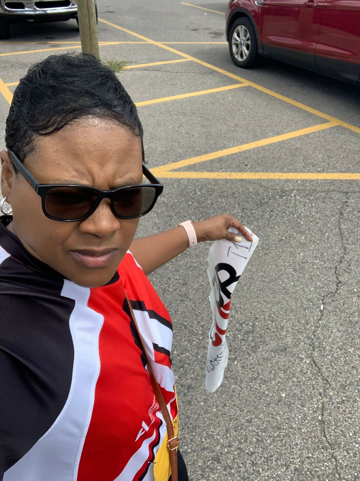 Person wearing sunglasses and a sports jersey, holding a rolled-up paper, standing in a parking lot with yellow lines.