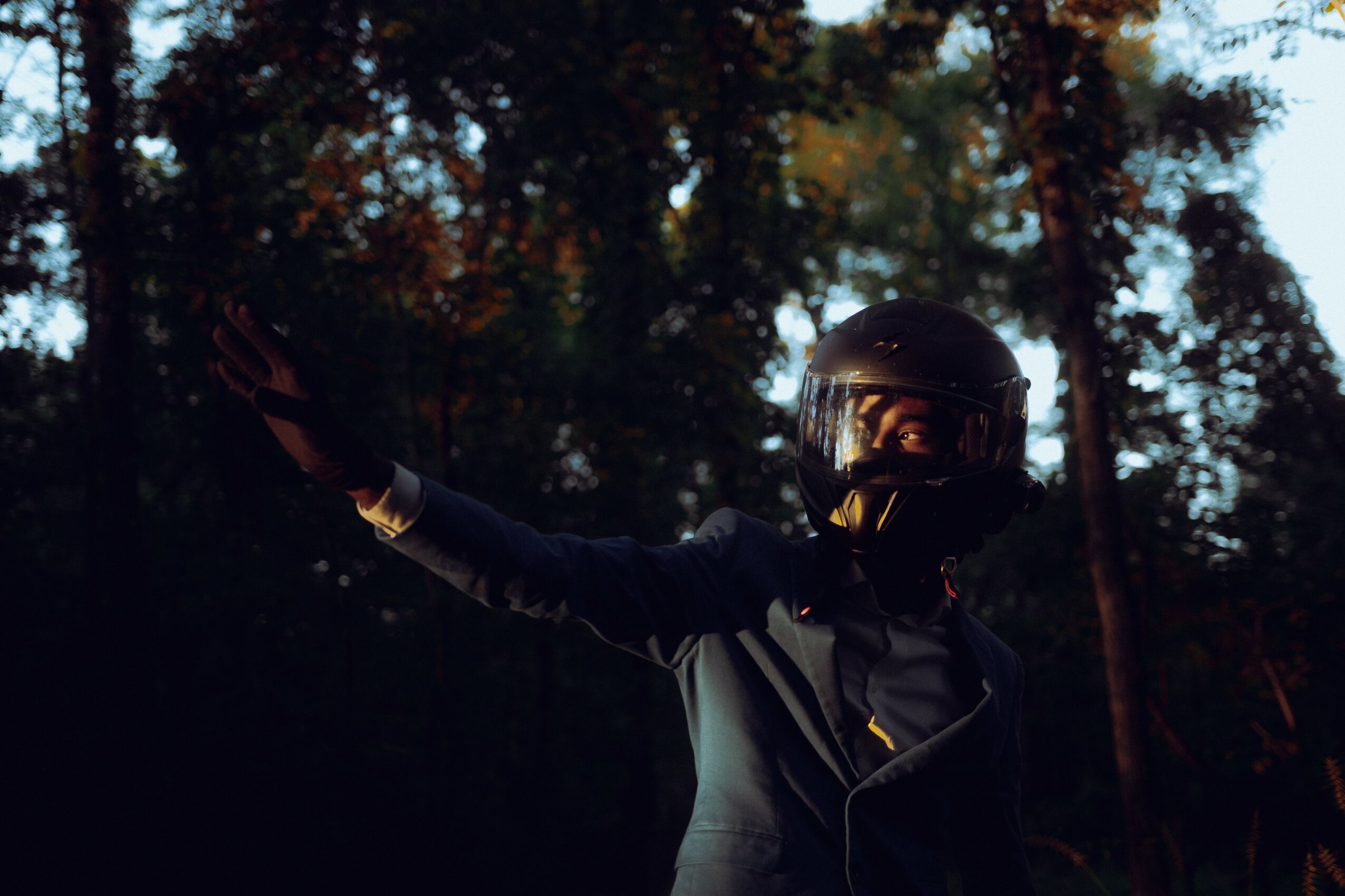Person wearing a helmet and jacket reaching out with an arm in a forest setting during sunset.
