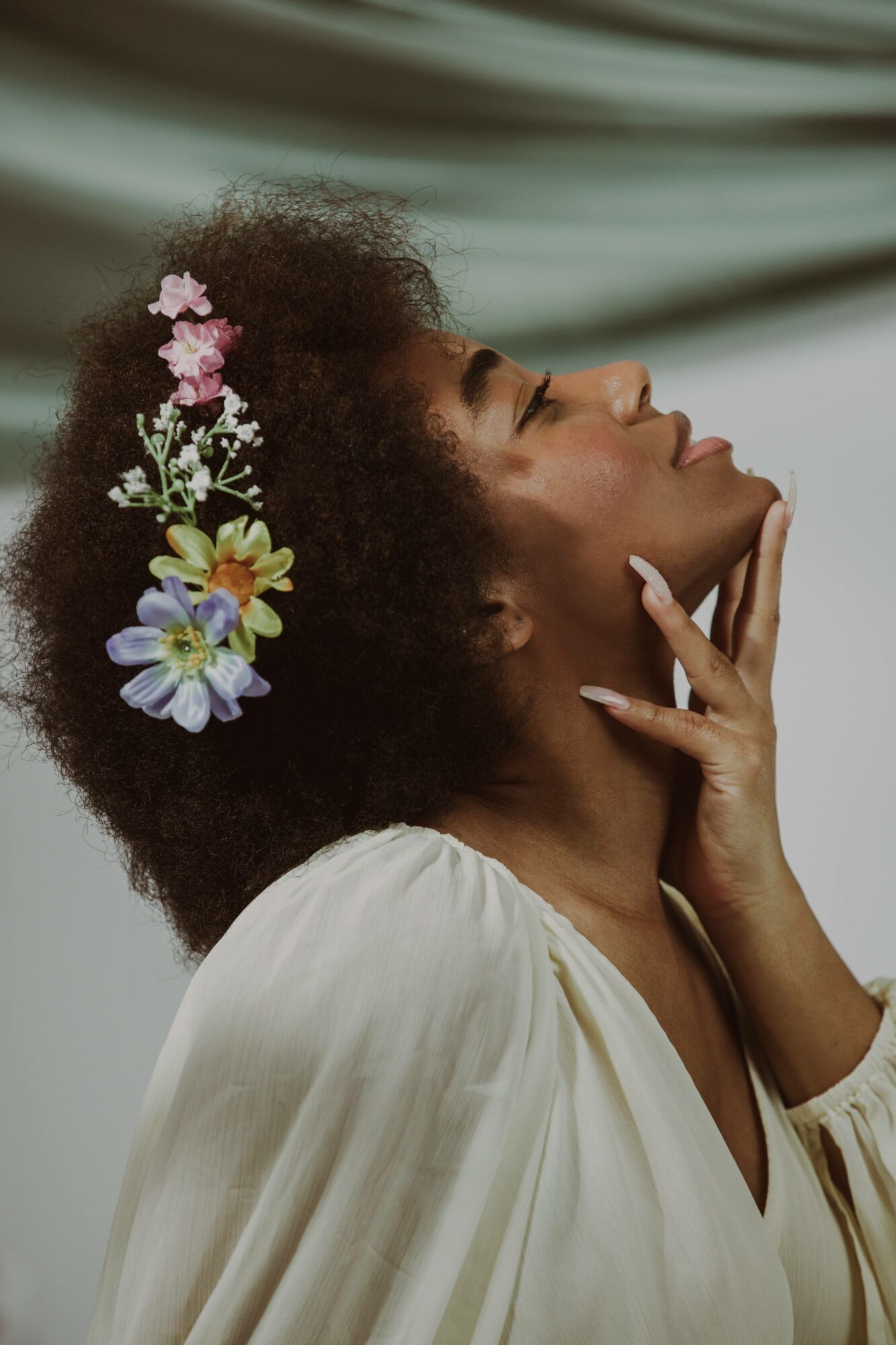Profile of a woman with curly hair and flowers, touching her face, wearing a white dress, against a soft background.
