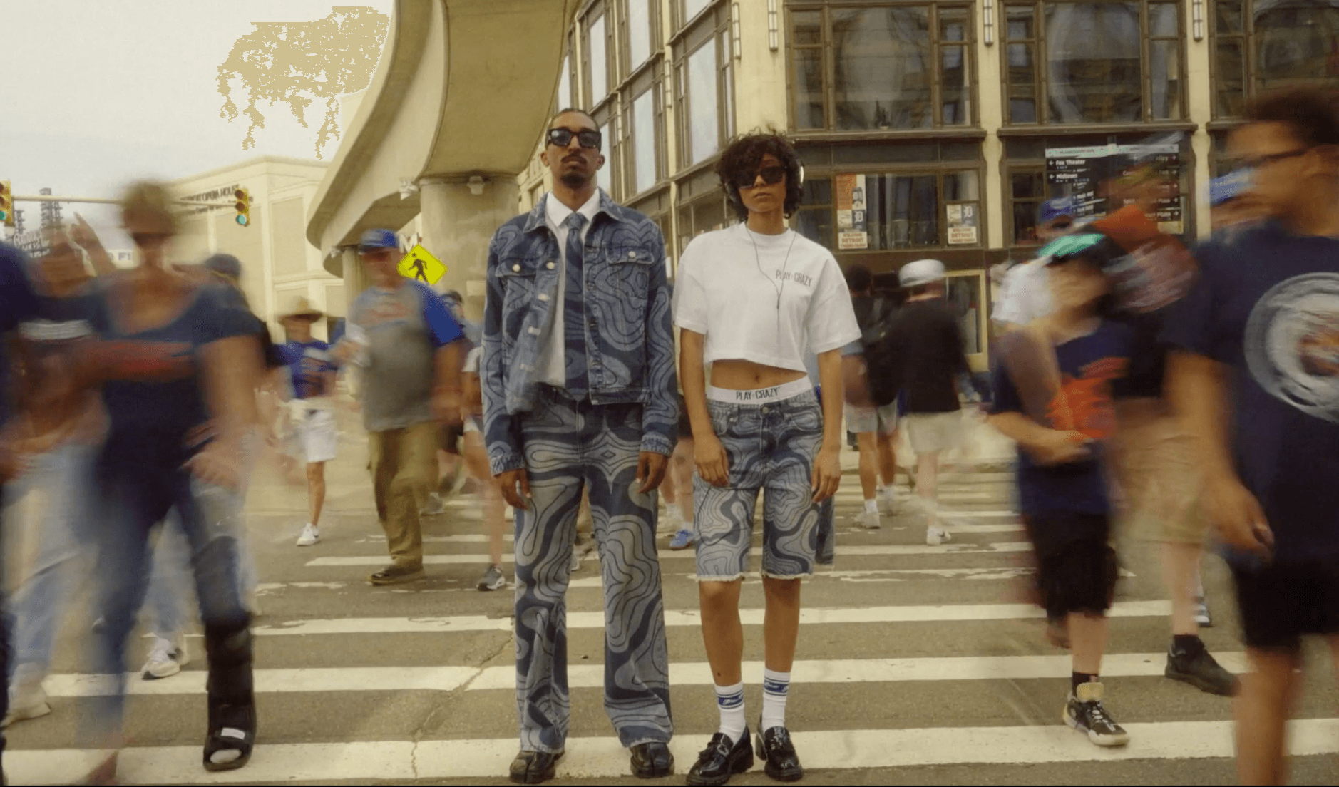 Two people stand on a crosswalk in a busy urban area with many pedestrians around, buildings, and an overpass in the background.