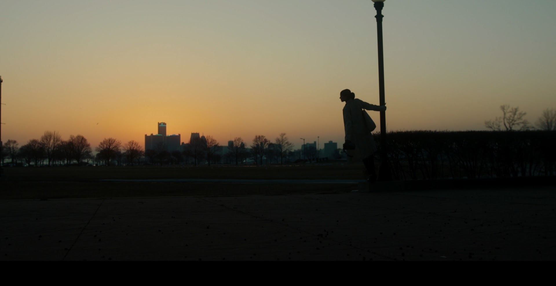 Silhouette of a person leaning on a lamppost during sunset, with a city skyline in the background.