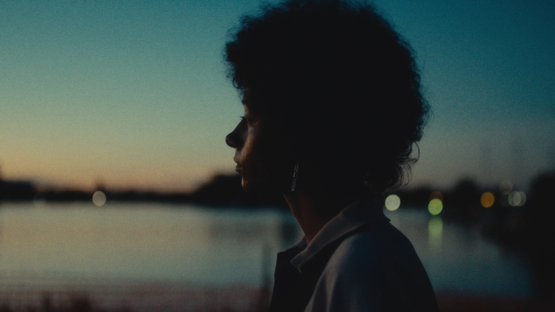 Silhouette of a person with curly hair against a sunset by water, with city lights in the distance.