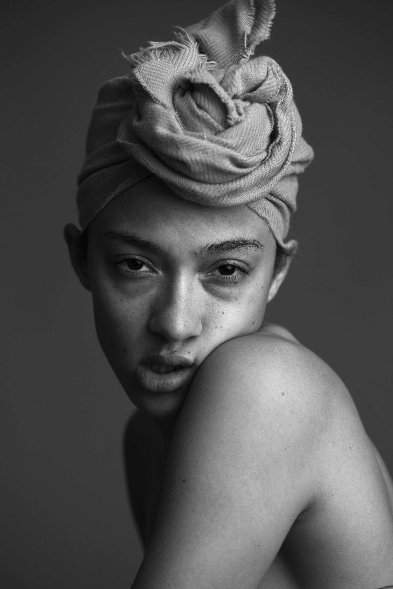 Young woman with head wrapped in cloth, looking directly at camera, shoulder visible, black and white photo.
