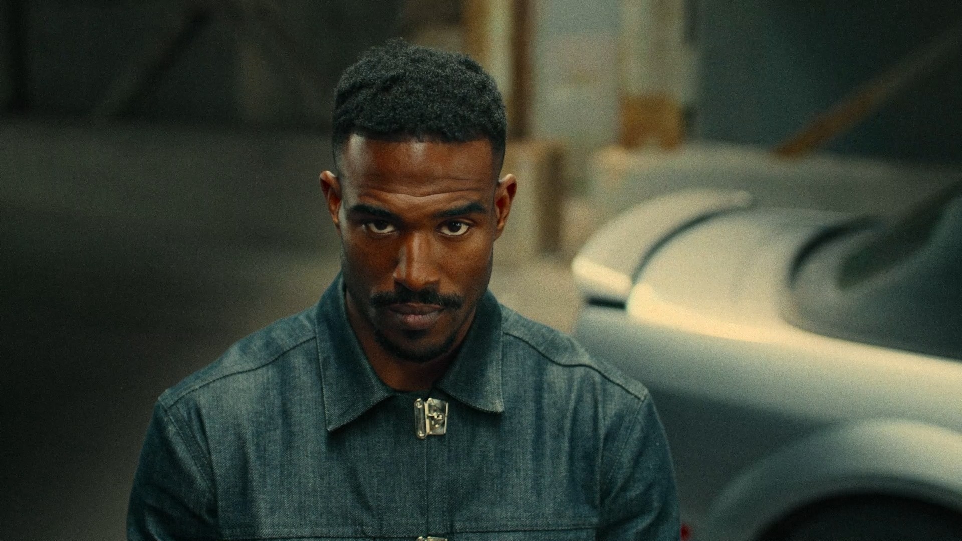 A man with short hair and a serious expression, wearing a denim jacket, standing outdoors near a car.