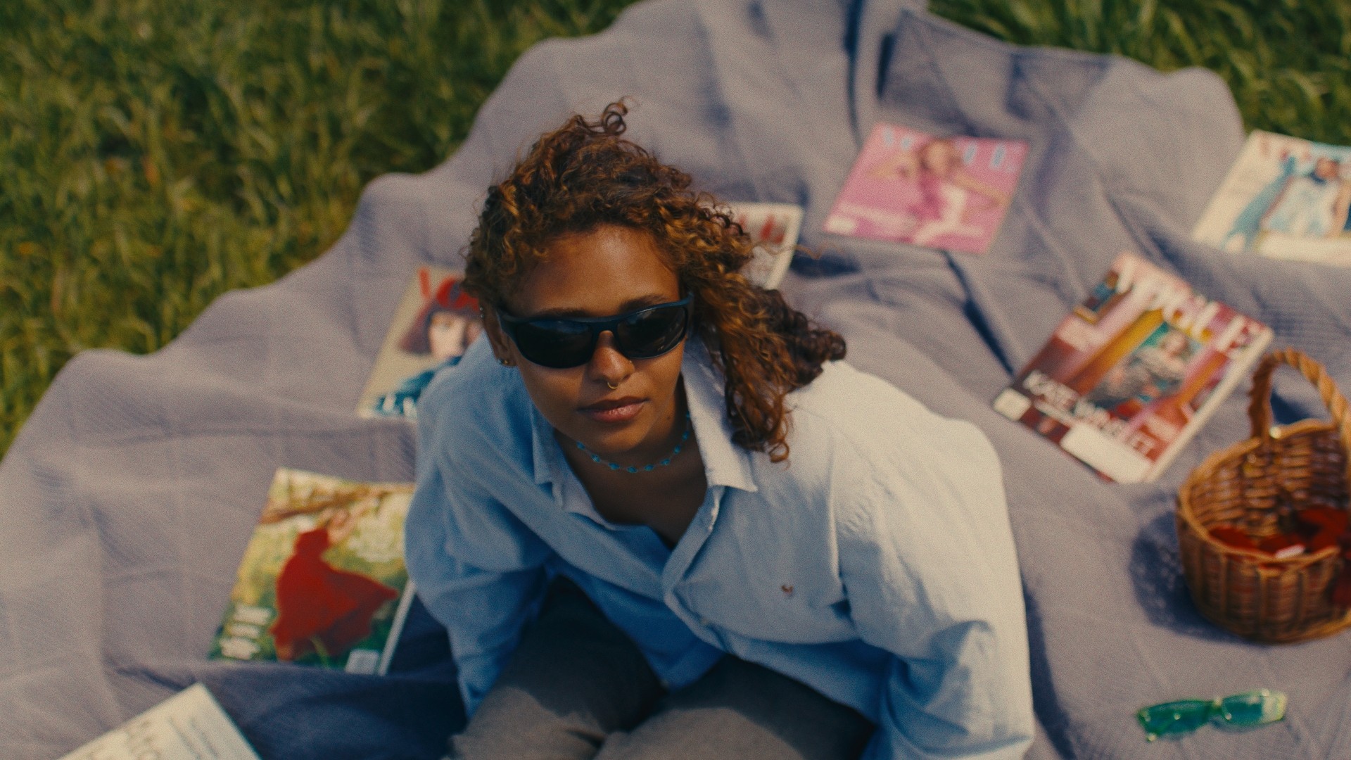 Woman with curly hair and sunglasses lying on a blanket outdoors, surrounded by books and a basket.