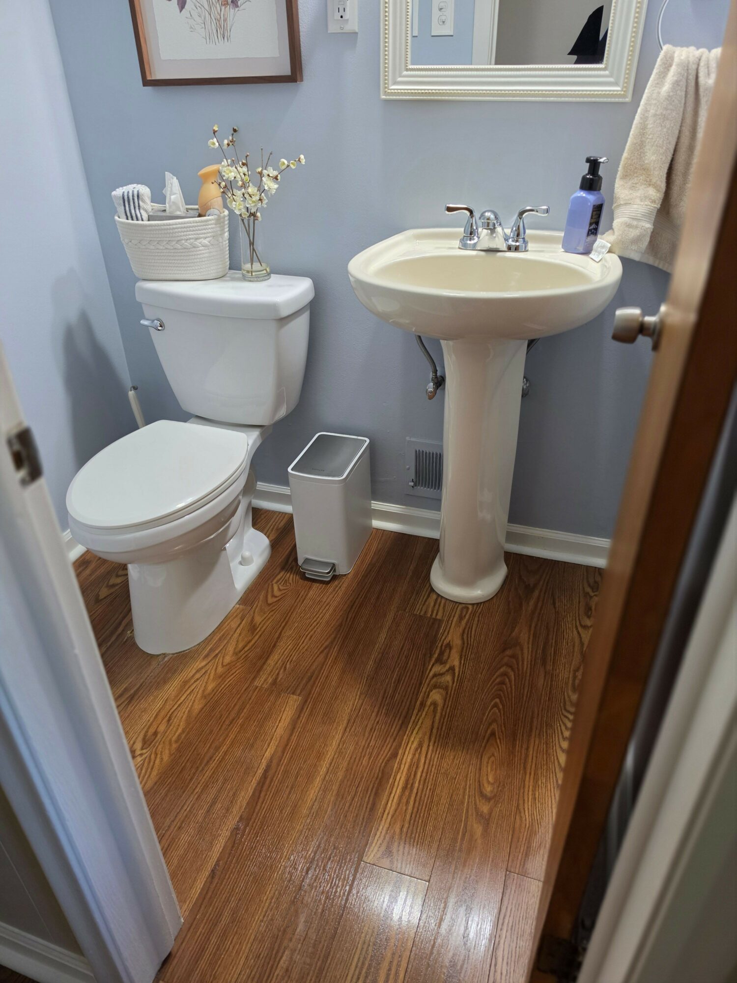 Bathroom with pedestal sink, toilet, small trash can, and wooden floor, viewed from doorway.
