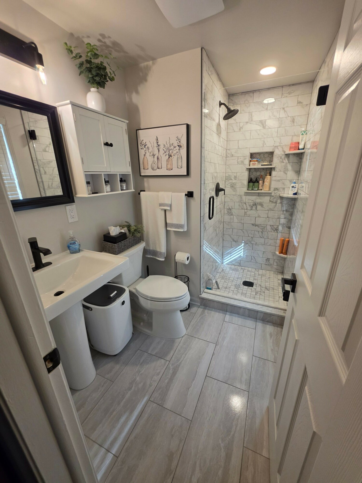 Bathroom with shower, toilet, sink, and storage cabinets, decorated in neutral tones with plants and framed art.