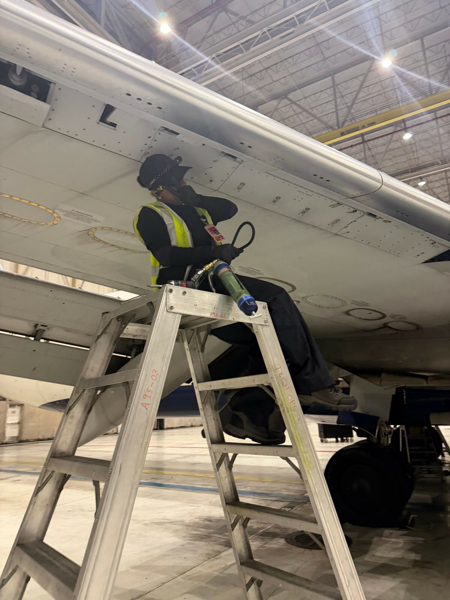 Person working on aircraft wing with ladder inside hangar, tools and equipment nearby.