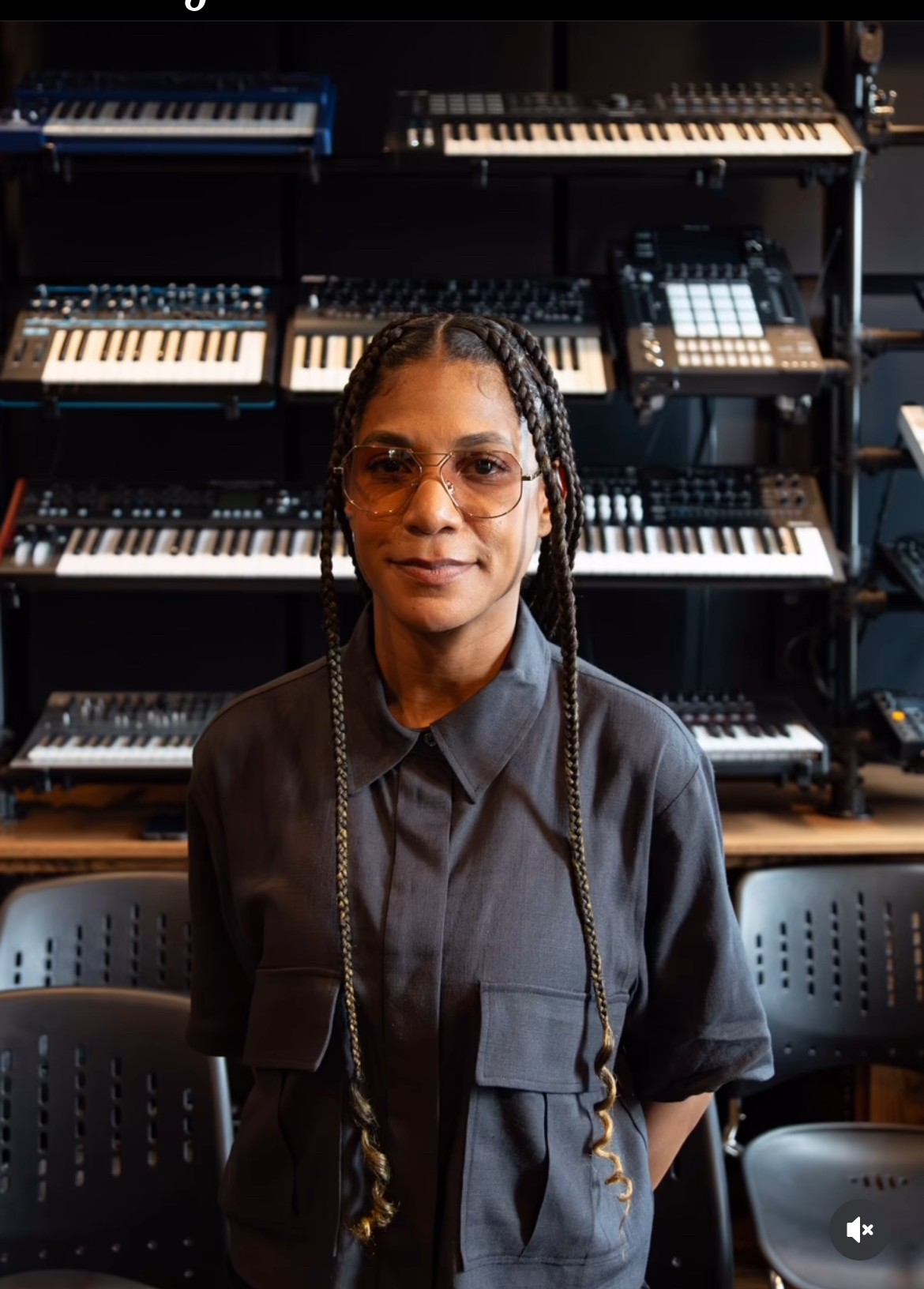 Person with braided hair and glasses standing in front of a wall of keyboards in a music studio.