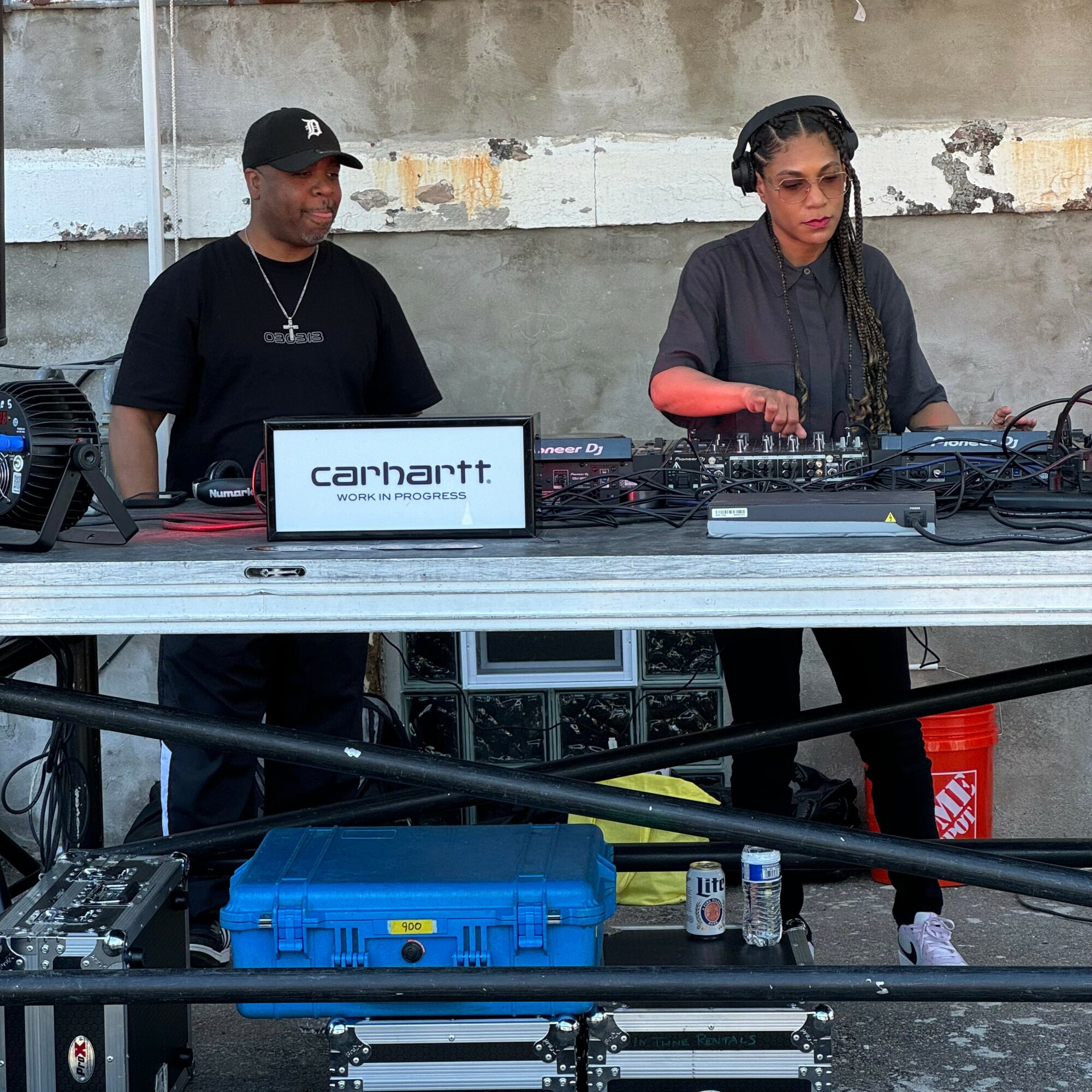 Two people DJing outdoors behind a table with equipment, a Carhartt sign, and a blue case, against a weathered wall.