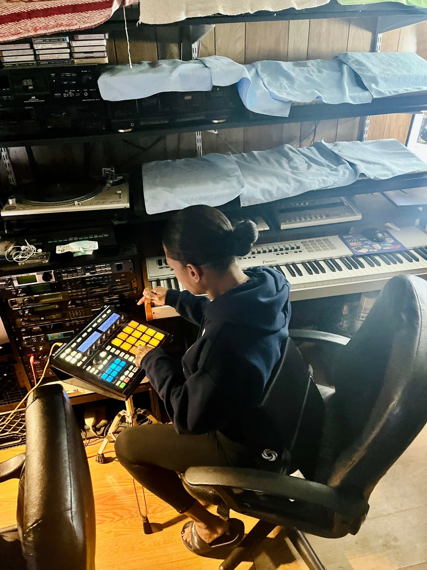 Person sitting on a chair using a music production device in a room with shelves of blue folded clothes and a keyboard, viewed from behind.