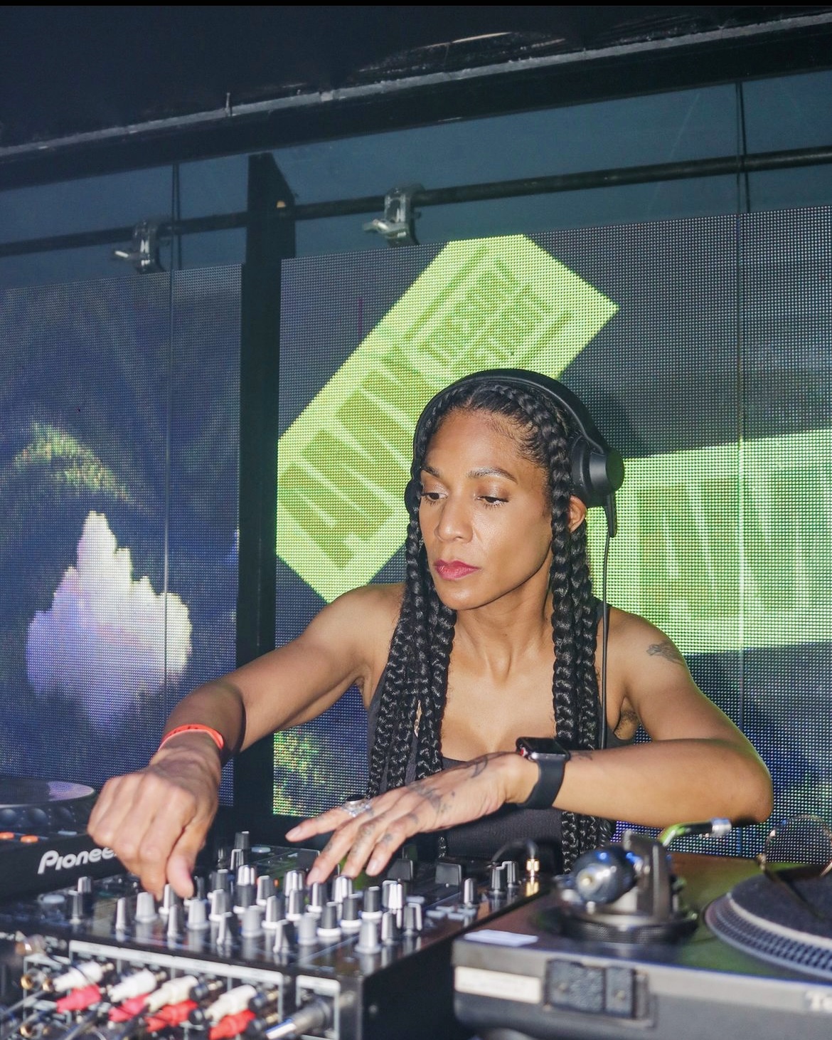 Female DJ with braided hair wearing headphones, operating DJ equipment in a studio with green and blue background