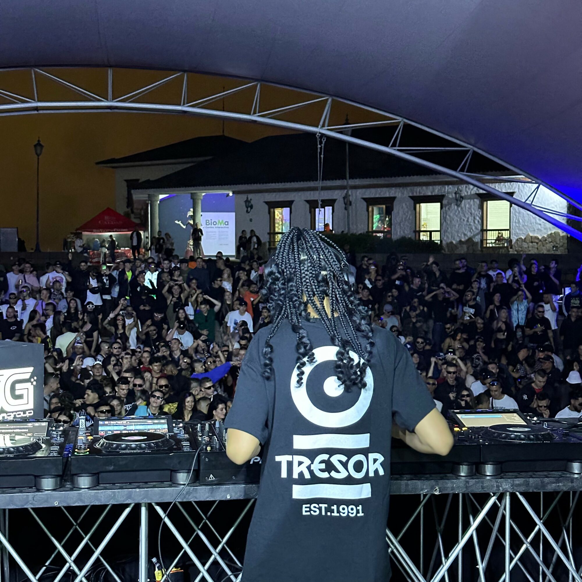DJ with braided hair performing at a crowded indoor event with a large audience and stage lighting.