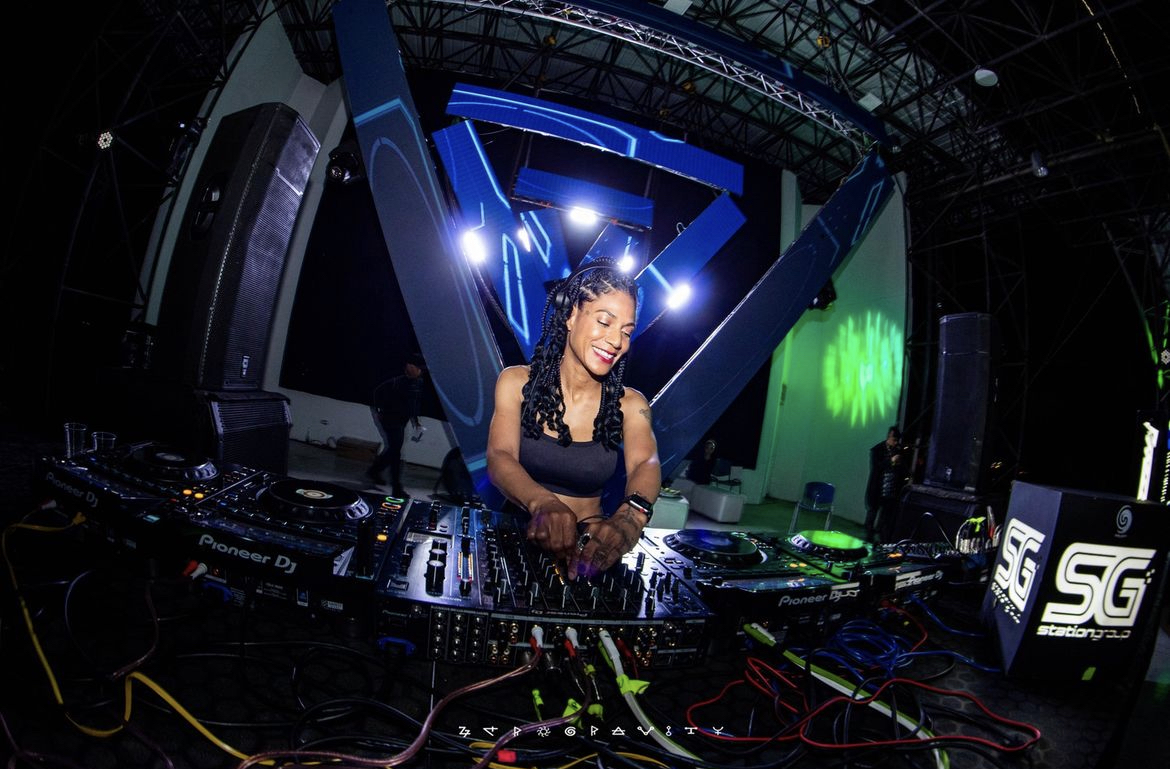 Female DJ with braided hair performing at a nightclub with colorful lights and DJ equipment.