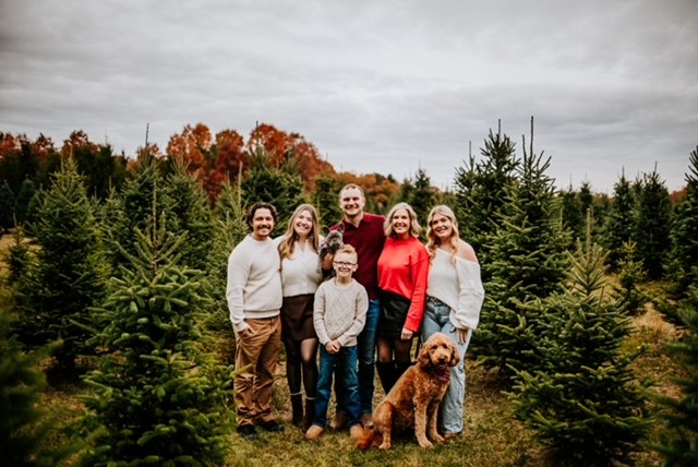Group of six people and a dog standing outdoors among trees, with autumn foliage and cloudy sky in background.