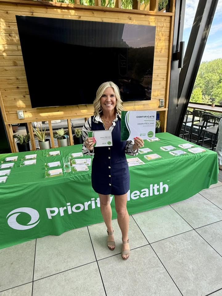 Woman holding a certificate and a green folder, standing in front of a table with a green tablecloth, outdoors.