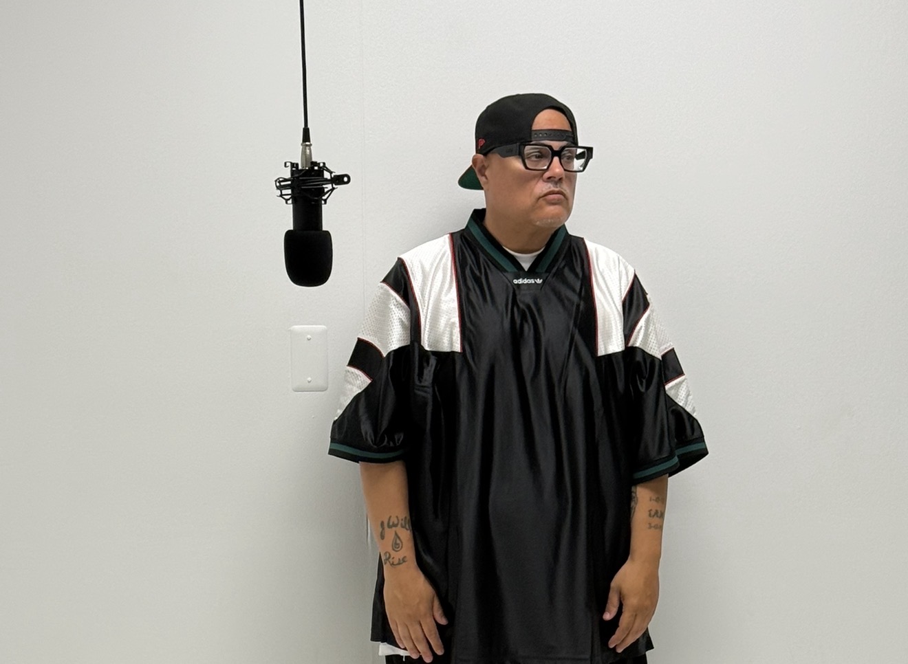 Person wearing glasses, a black cap backwards, and a black oversized shirt, standing against a plain white wall with a microphone hanging nearby.