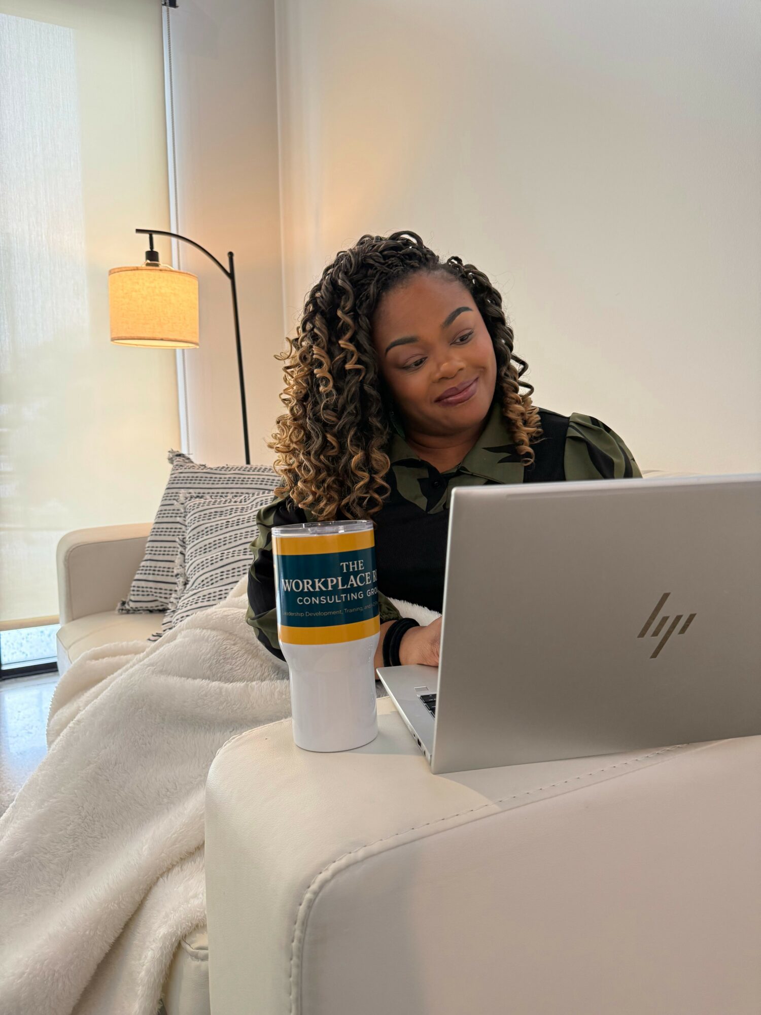 Woman working on laptop with coffee cup nearby in a cozy room, smiling and looking at screen.
