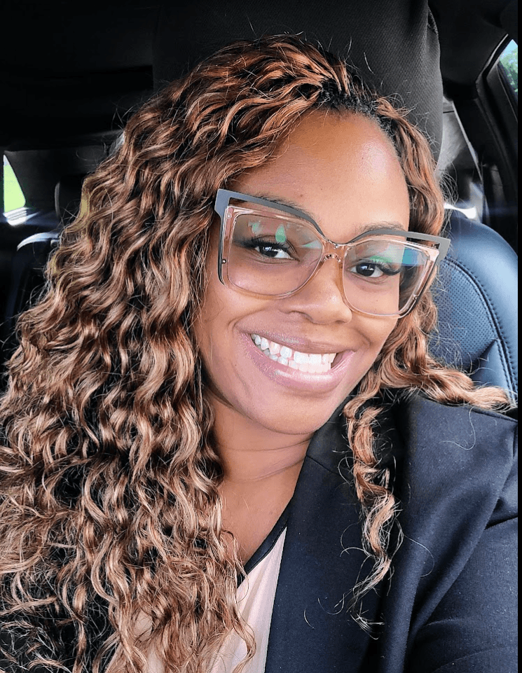 Woman with curly hair wearing glasses and smiling, sitting in a car seat.