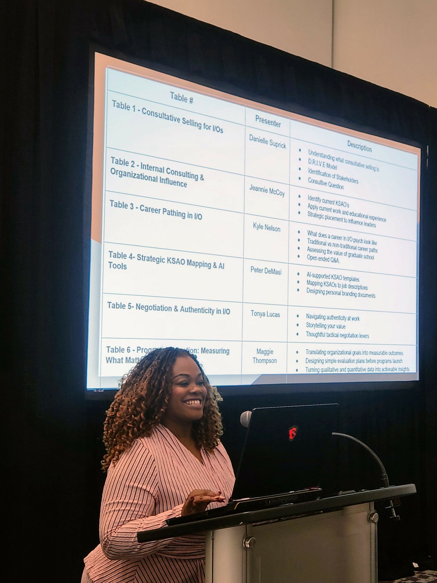 Smiling woman with curly hair standing at a podium in front of a presentation screen displaying a table with course titles and descriptions.