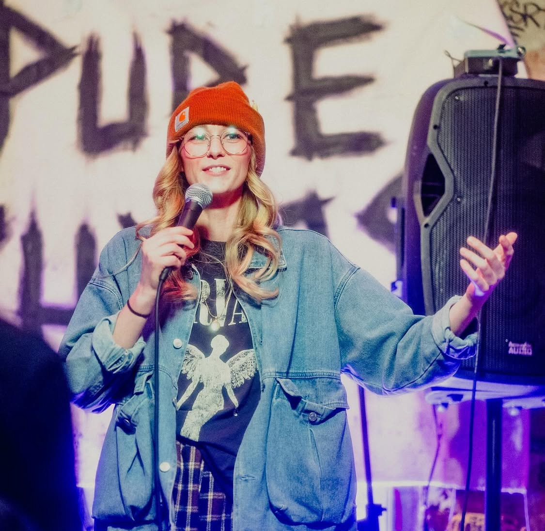 Young woman with glasses and a red beanie holding a microphone on stage, smiling, with a speaker nearby.