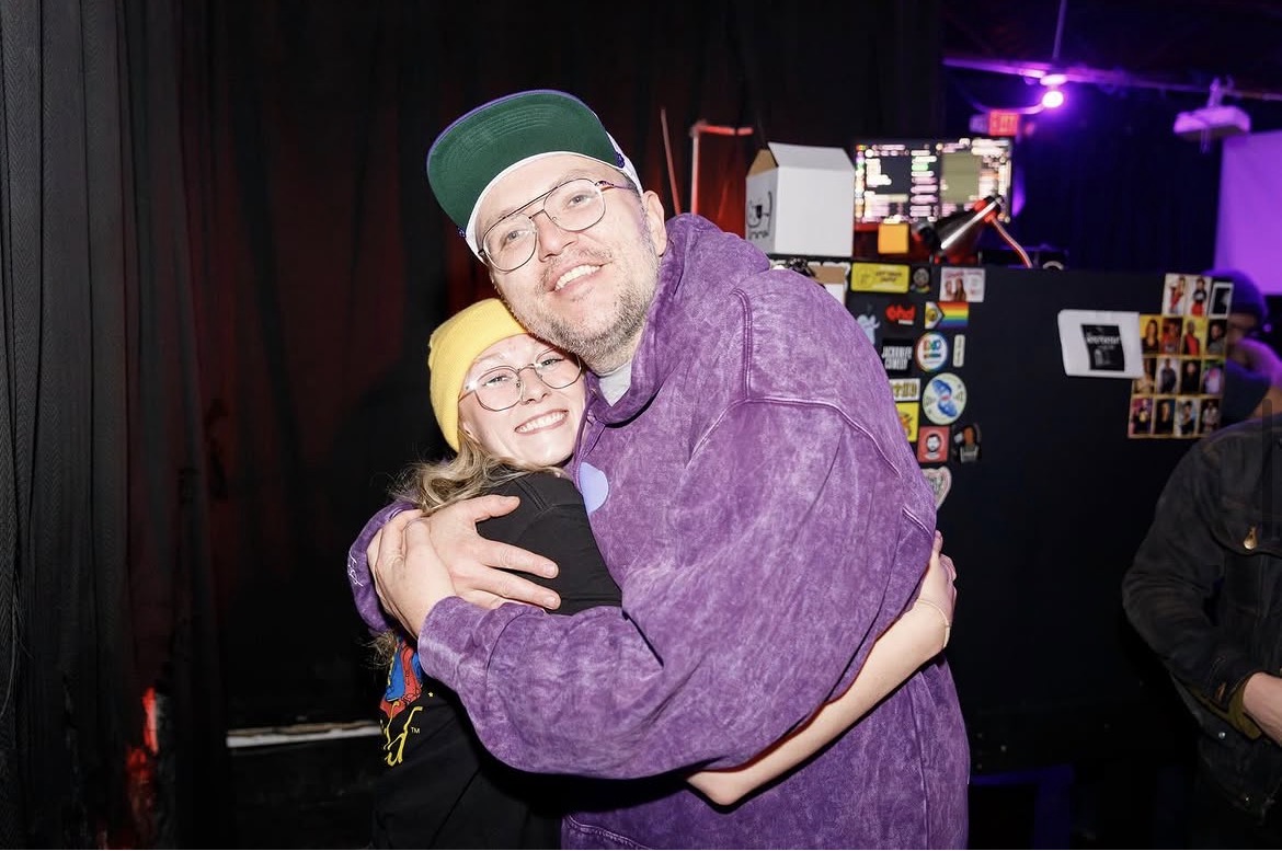 Two people hugging, smiling, in a dimly lit room with colorful lights and equipment in the background.