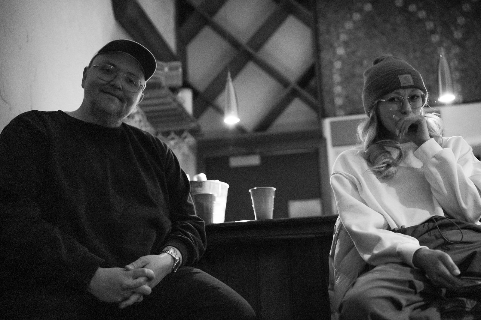 Two people sitting at a bar, one smiling and the other wearing a beanie and glasses, in a dimly lit setting.