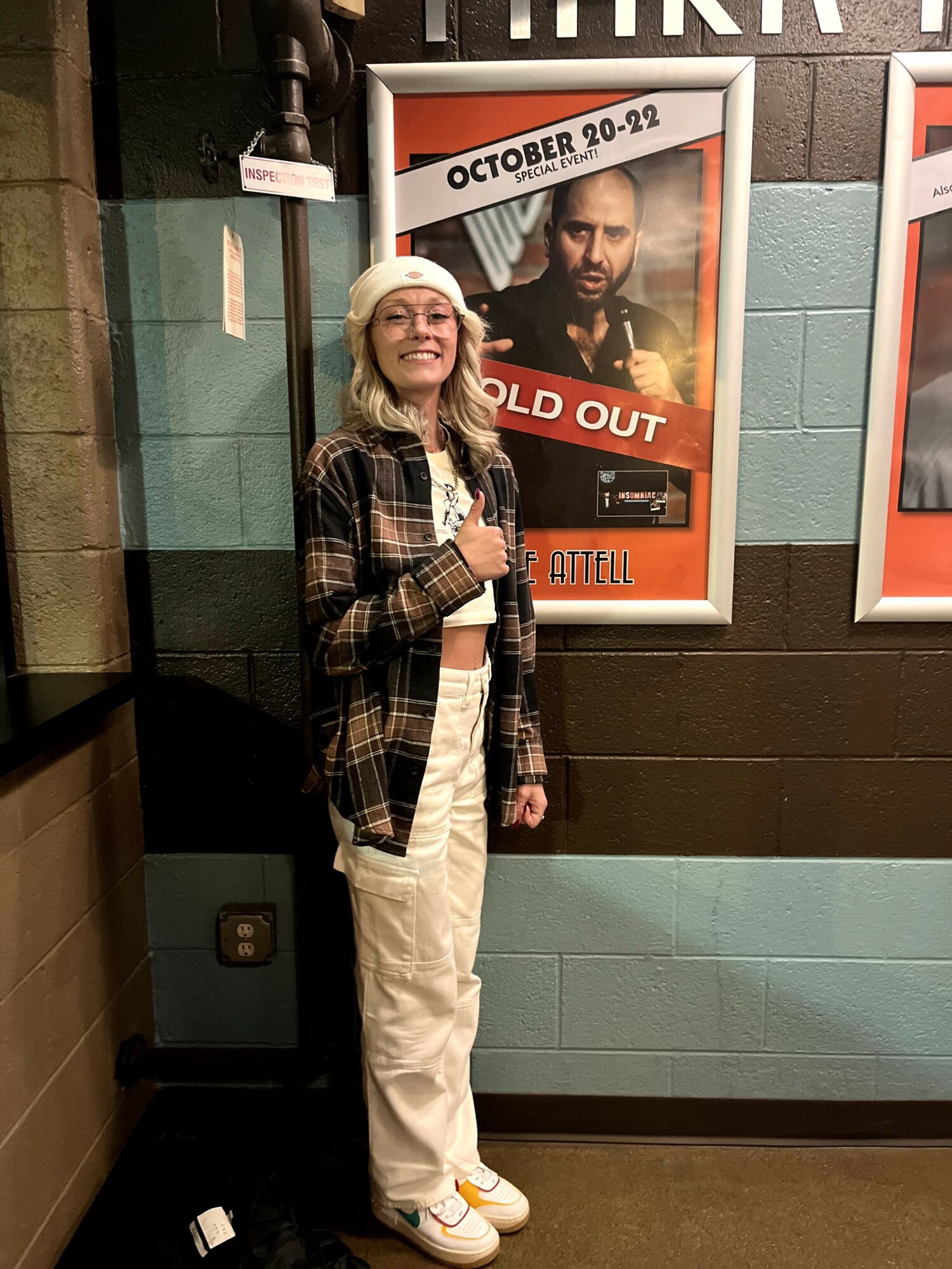 Young woman with glasses and a Santa hat stands in front of a poster, holding a drink, smiling, wearing a plaid shirt and white pants.