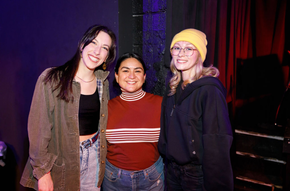 Three women standing together, smiling, in a dimly lit indoor setting with dark background and red curtains.