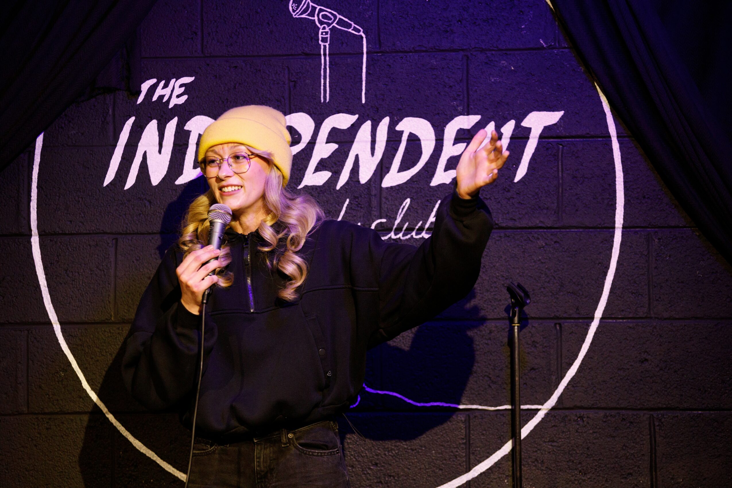 Woman with glasses and yellow hat speaking into a microphone on stage, with a logo behind her.