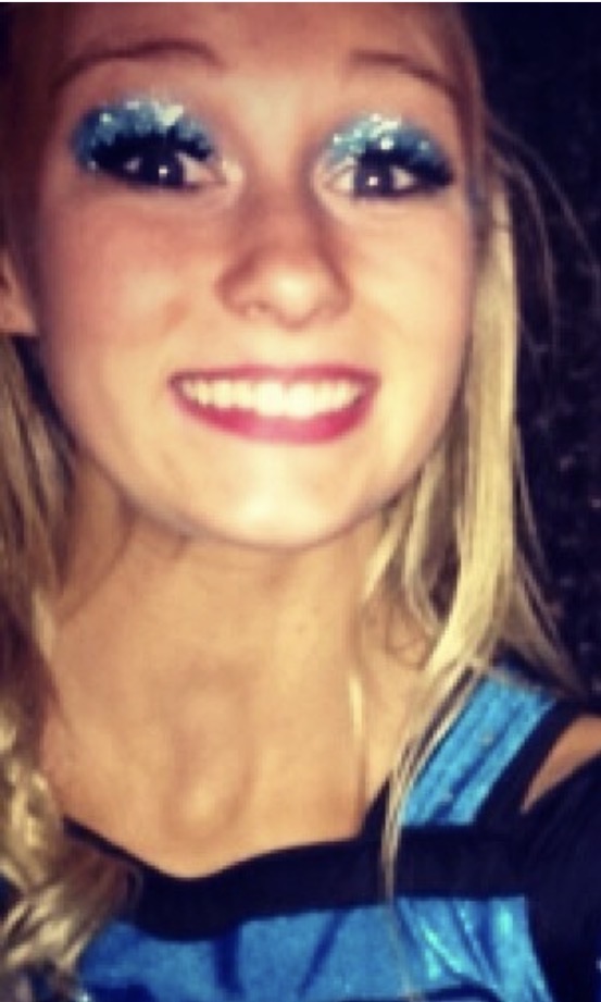 Close-up of a smiling woman with blue glittery eye makeup and blonde hair, wearing a blue and black top.