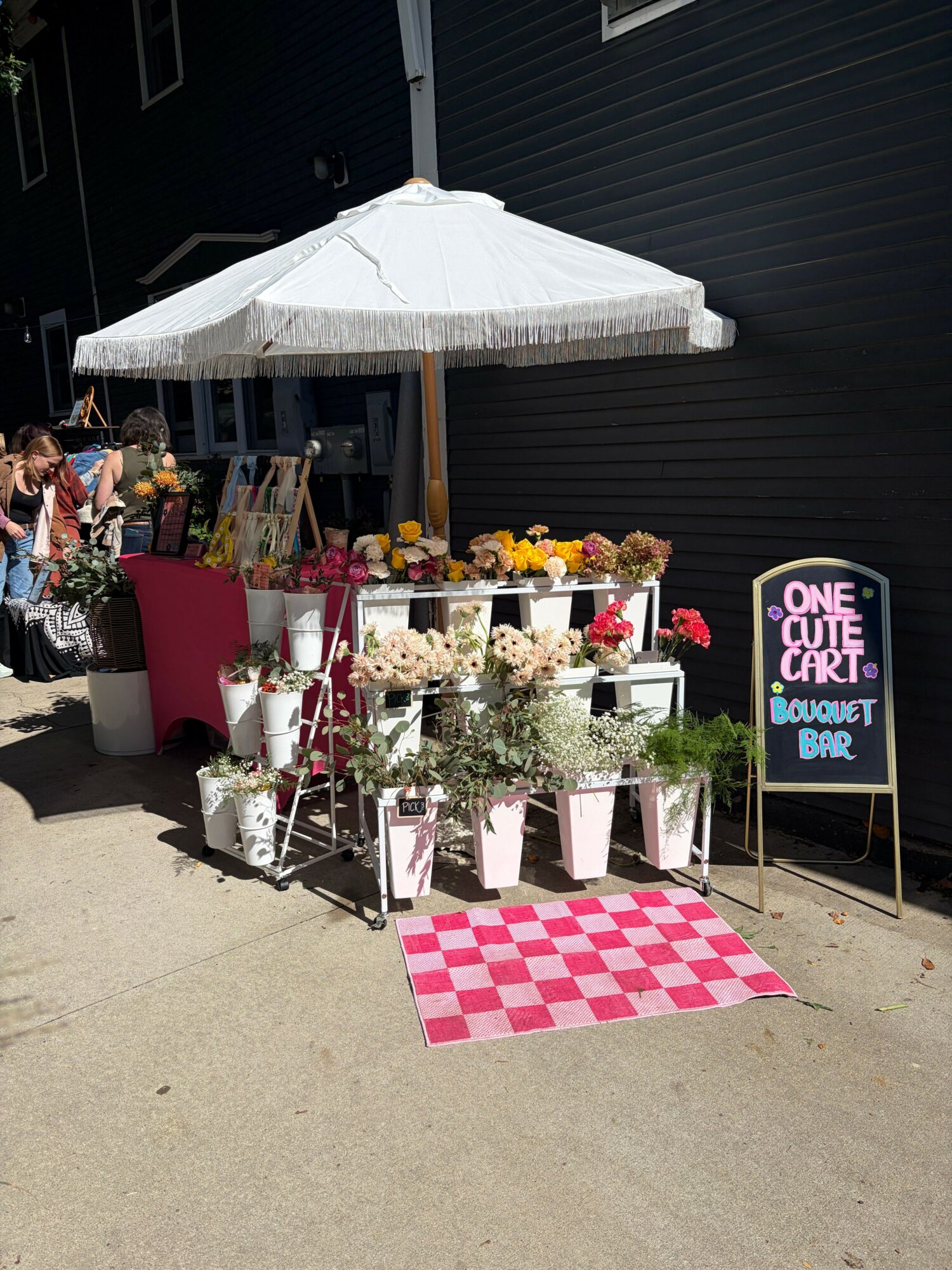 Outdoor flower display with various plants and flowers, a pink table, and a sign reading 'ONE OF A KIND' on a black wall.