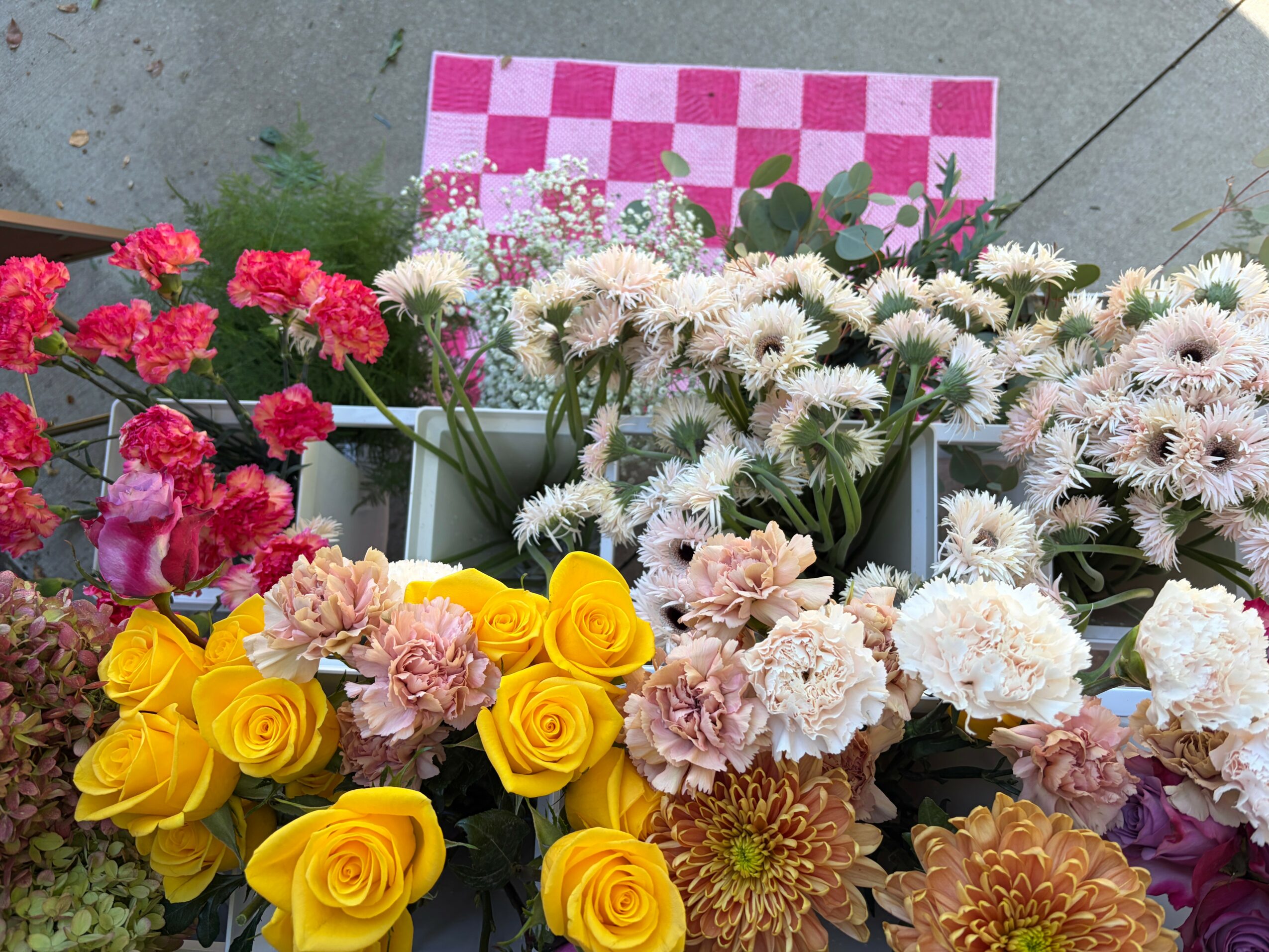 Various colorful flowers including pink, white, yellow, and purple blooms arranged together, with a checkered pink and white cloth in the background.