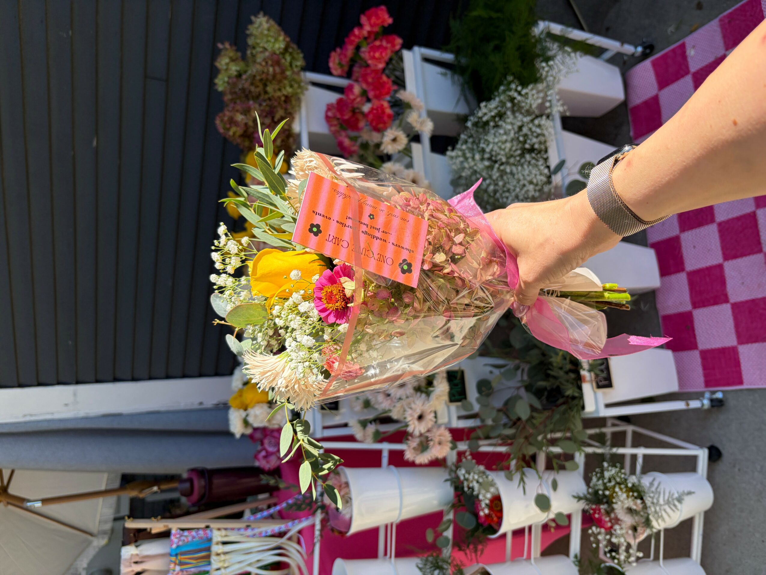 Person holding a bouquet of colorful flowers with a pink card, outdoors with plants and furniture in background.